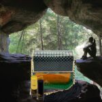 A man sitting in front of a cave with a bottle and glass.