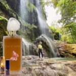 A person standing in front of a waterfall with a bottle of perfume.