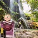 A person standing in front of a waterfall with a bottle of perfume.