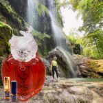 A person standing in front of a waterfall with a bottle of perfume.