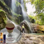 A person standing in front of a waterfall with a bottle and some oils.